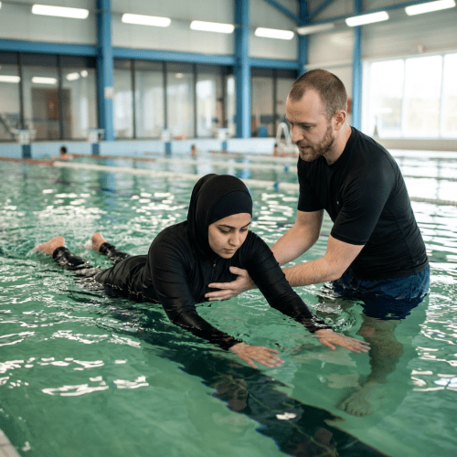 Woman in black full-body swimsuit practicing swimming in indoor pool with instructor's assistance